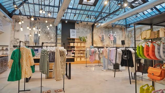 Intérieur d'une boutique moderne avec des vêtements suspendus sur des supports, des étagères en bois et un plafond en verre, offrant une ambiance lumineuse et aérée.