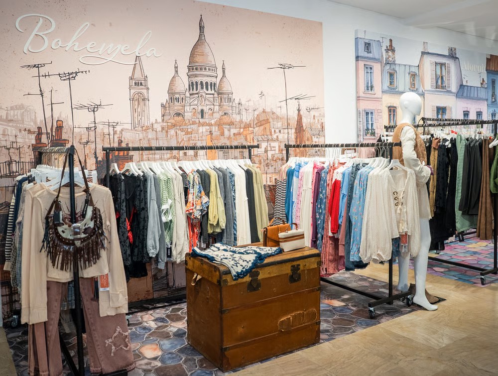 Vue d'un magasin de vêtements avec une ambiance boho, présentée sur un fond illustré de la ville de Paris, mettant en valeur différents styles de mode accrochés sur des portants.