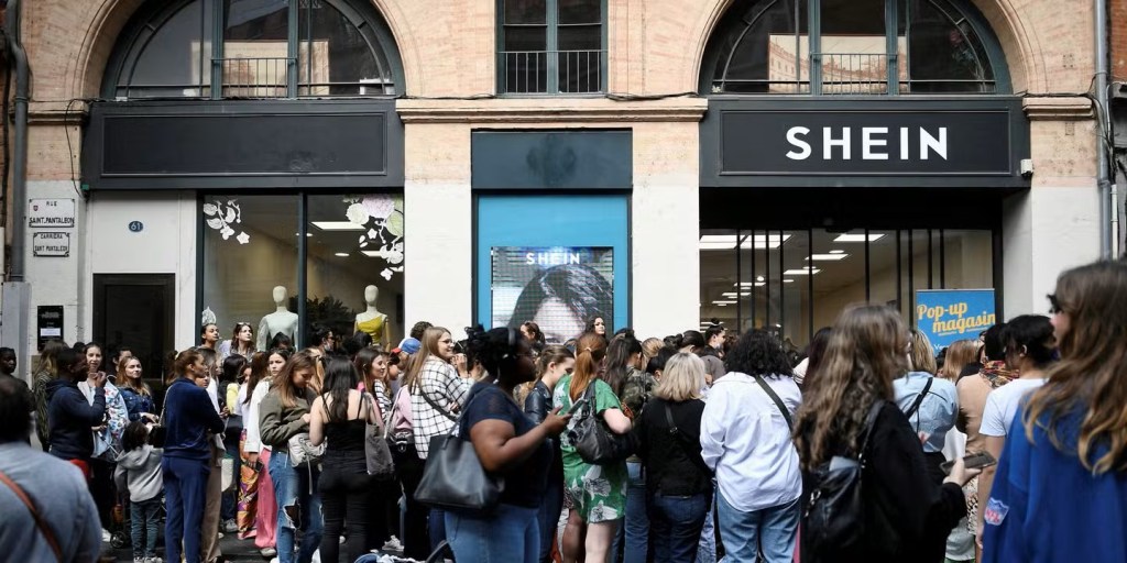 Une foule de personnes attend devant la boutique SHEIN, avec des mannequins visibles à l'intérieur et des panneaux d'affichage à l'extérieur.